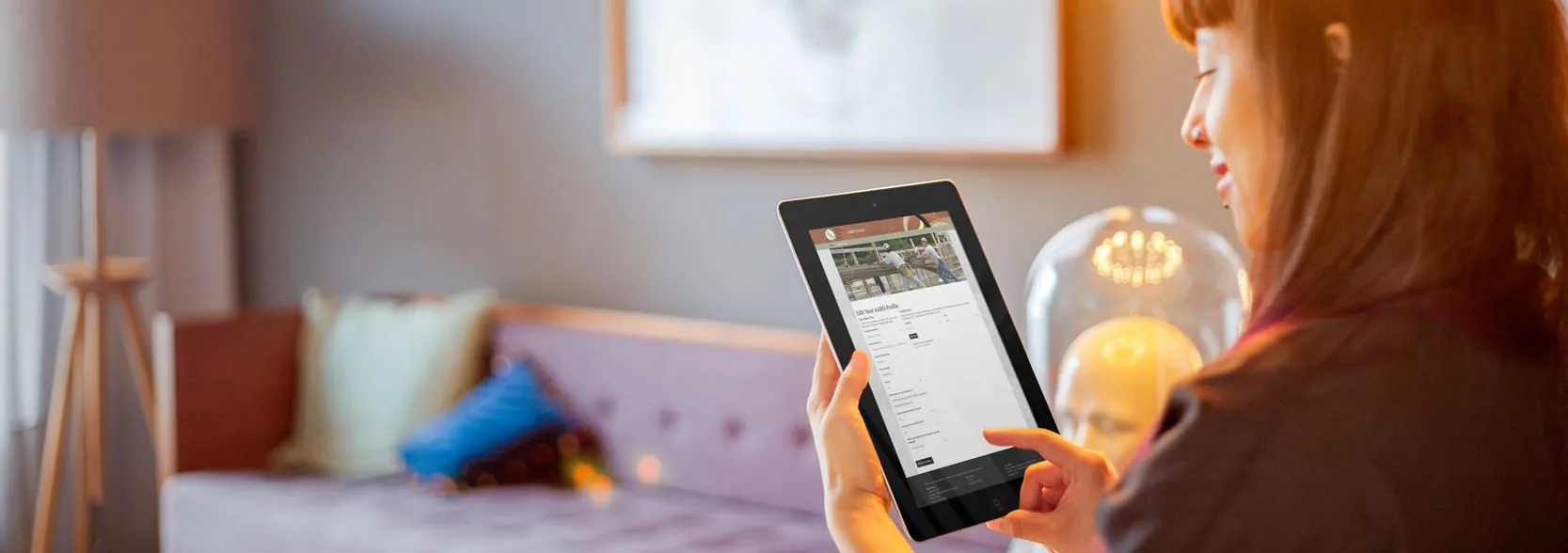 A woman on a sofa holding an iPad open to the AABO portal's Edit Your AABO Profile page