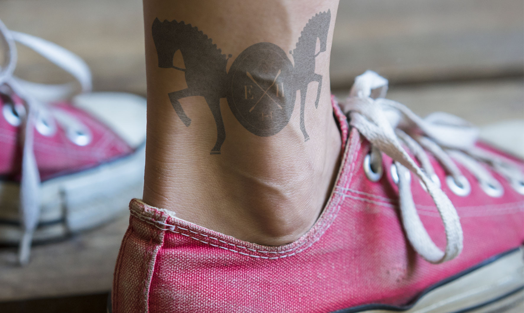 Close-up of an ankle tattoo of two horses around an "EH" emblem, above a red canvas sneaker