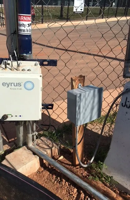A DCS enclosure bolted to a pole beside a chain-link fence on a building site, with a grey junction box on a post next to it