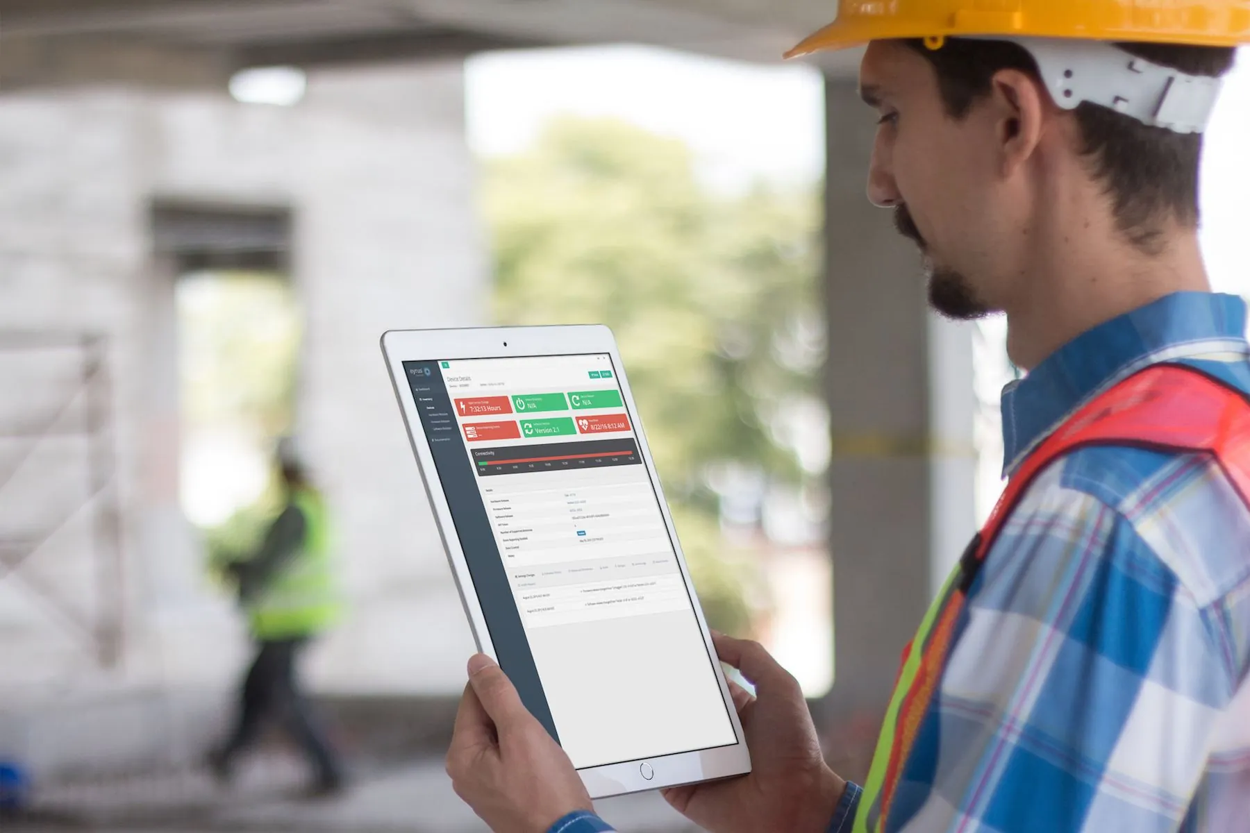 A construction worker in a hard hat and safety vest viewing a device dashboard on an iPad at a building site