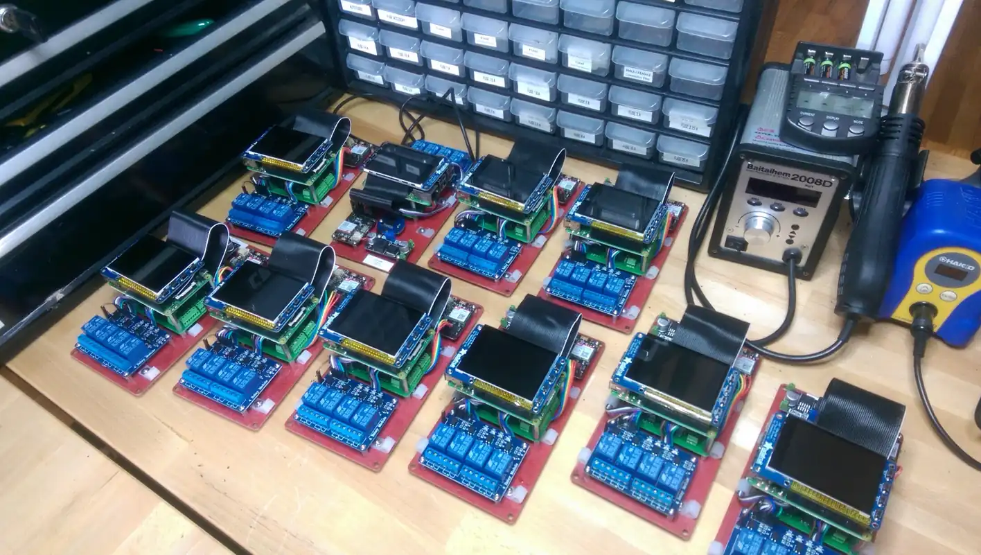 Ten DCS control boards laid out on a workbench, each a Raspberry Pi with a small screen above a bank of blue relays on a red baseplate, beside a parts drawer and a hot-air rework station