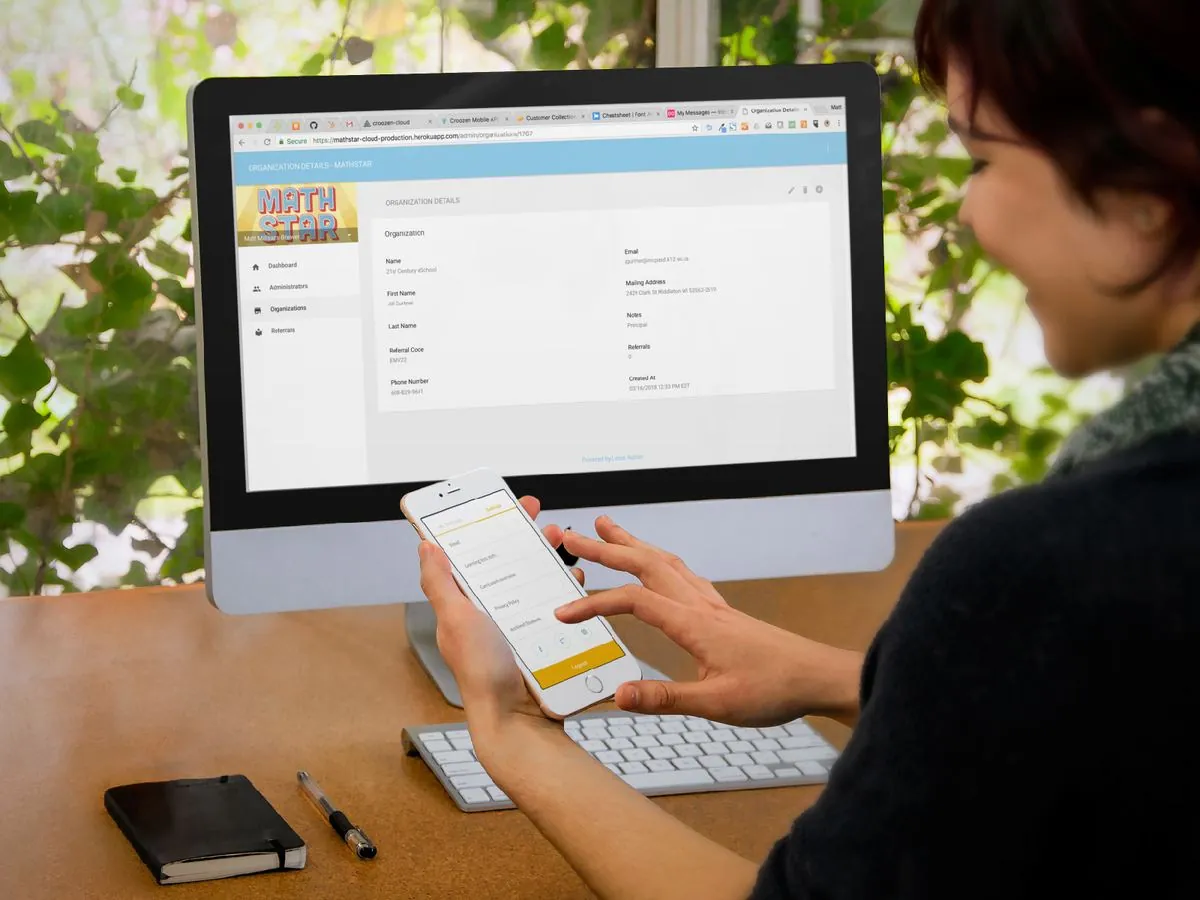 A woman using a phone at a desk in front of a monitor showing the Math Star back office's Organization Details page