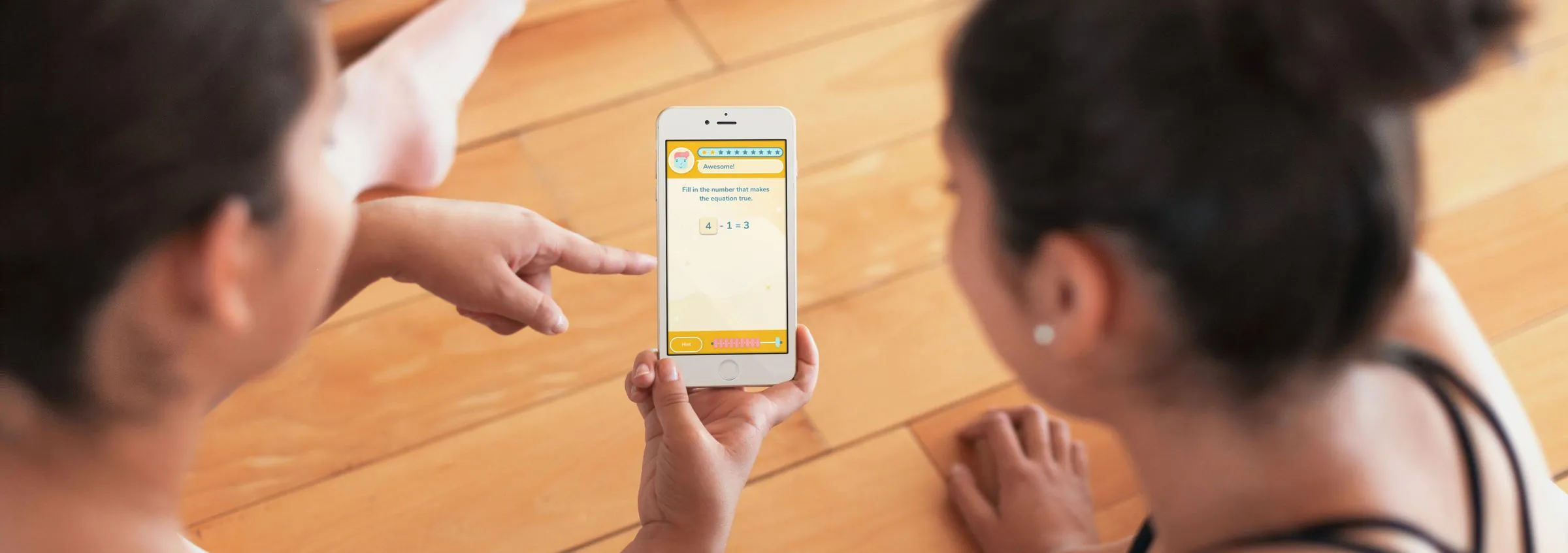 Two people sitting on a wooden floor looking at a phone showing a Math Star fill-in-the-number equation