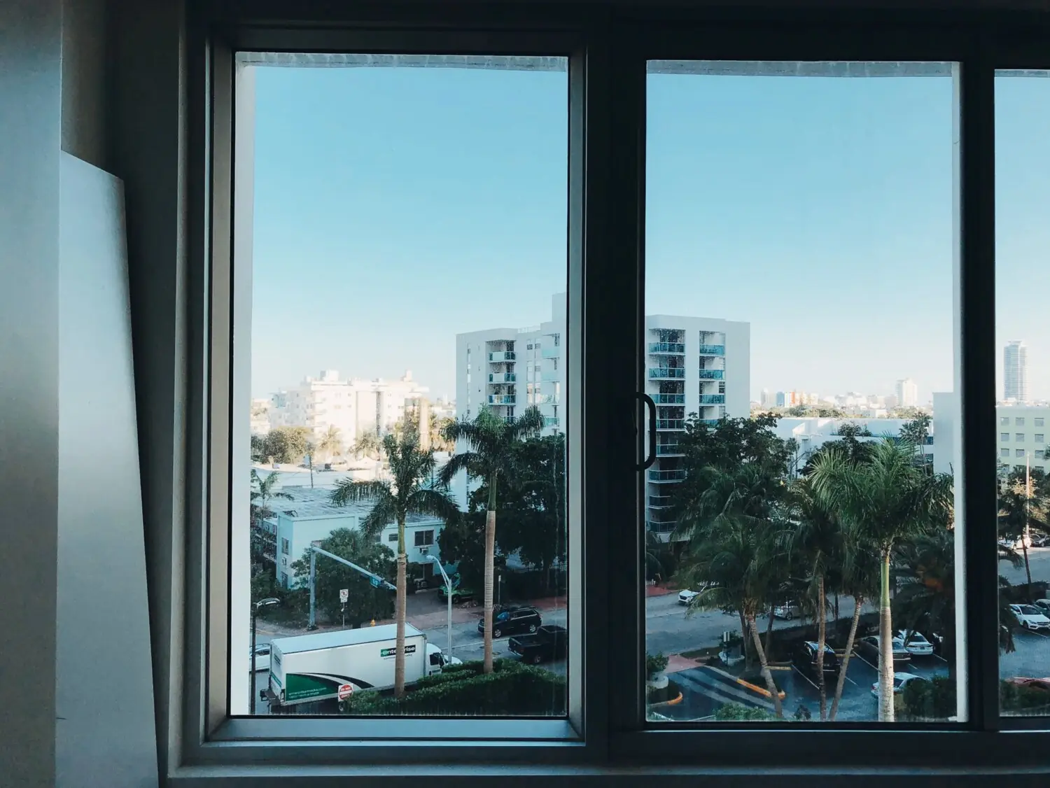 A photo taken through a wide window of two panes, looking out over palm trees and apartment blocks under a clear sky.
