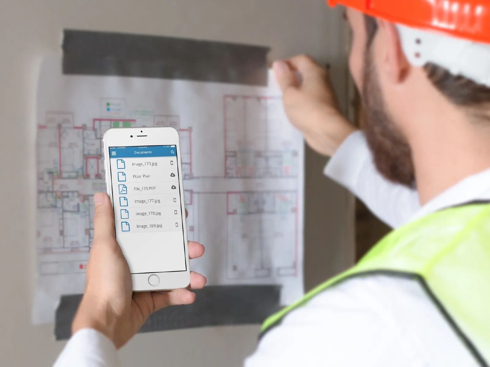A worker in a hard hat and safety vest holds a phone showing Project Boss documents in front of a floor plan