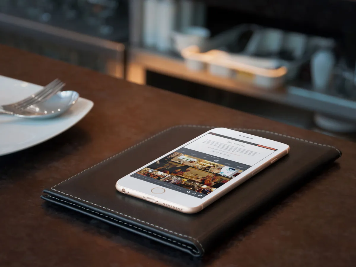 A phone lying on a leather check folder at a restaurant table, showing the Tasting Collective Our Mission page