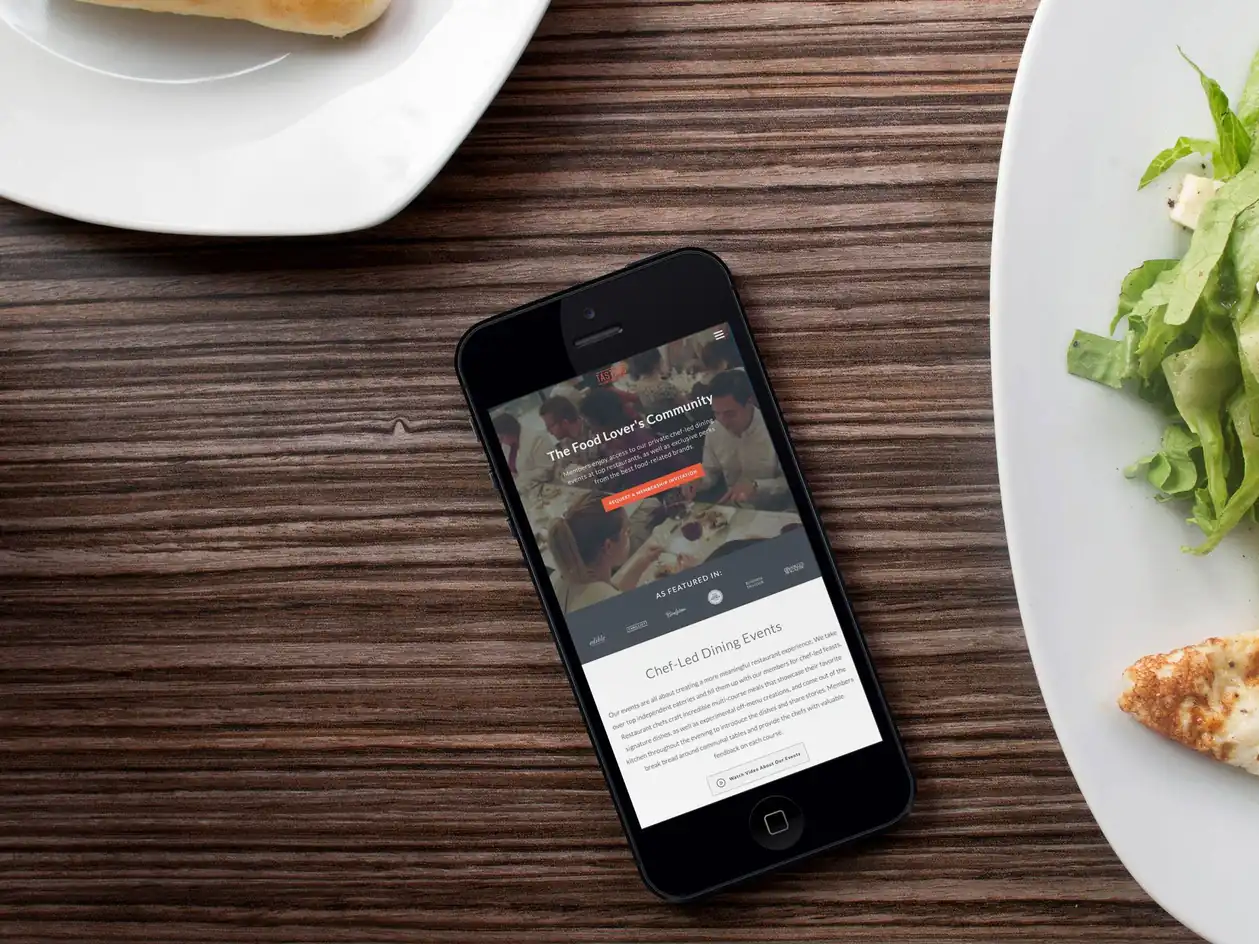 A phone on a wooden table between plates of food, showing the Tasting Collective site with the heading The Food Lover's Community