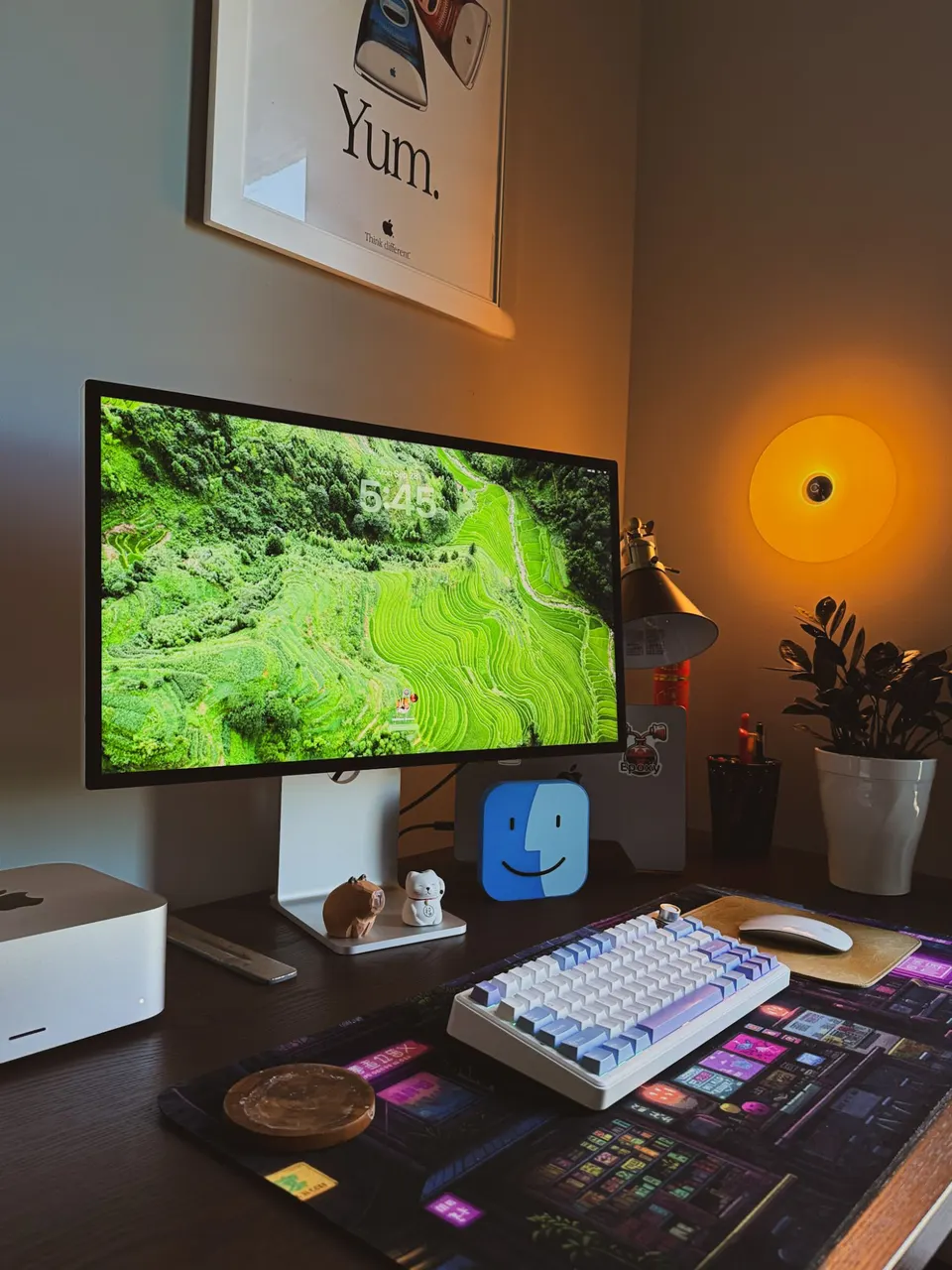 Matt's desk in the evening: a big display, a white and lilac keyboard, a small wooden capybara, and a framed “Yum.” poster lit by an orange lamp