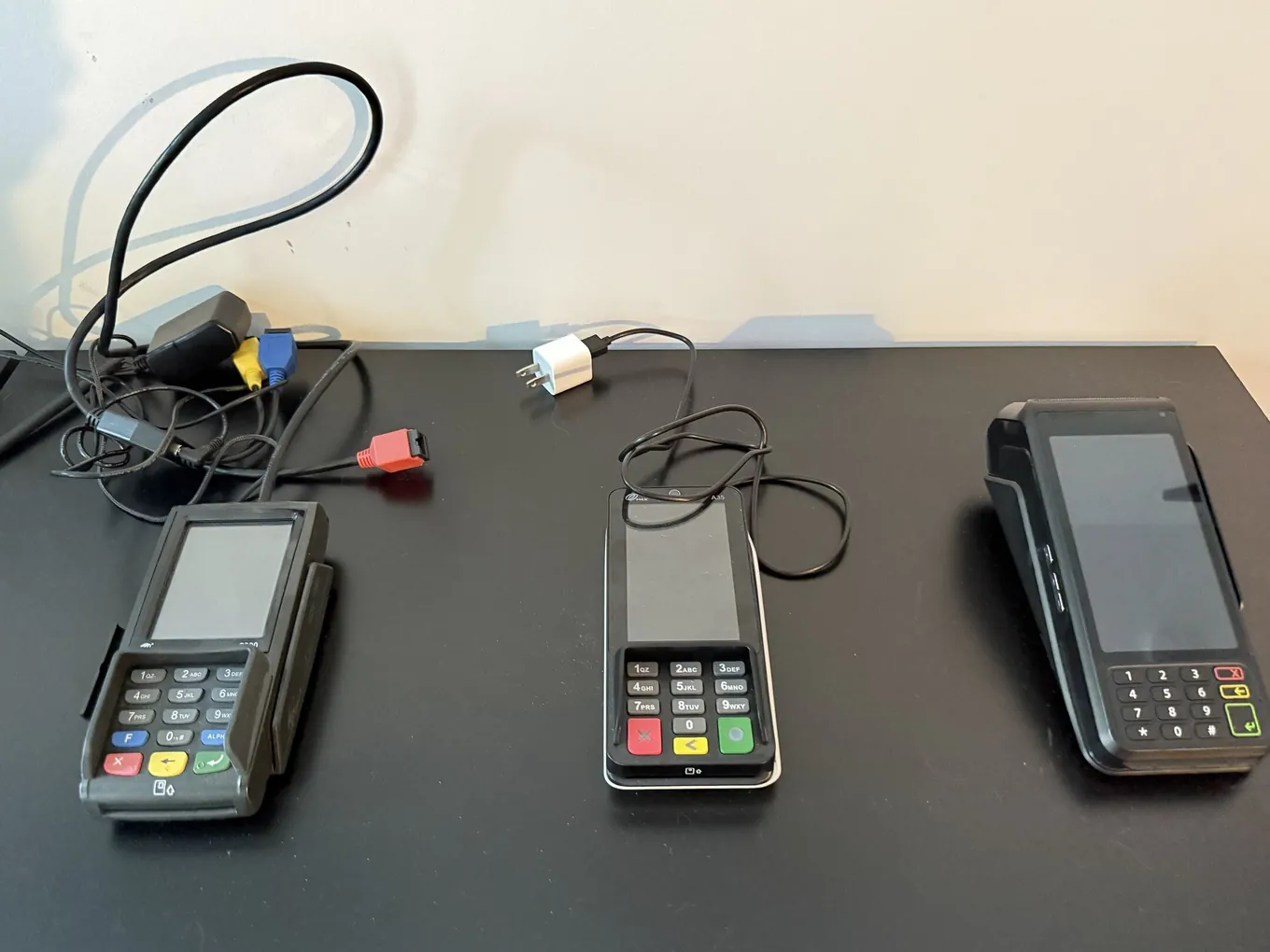 Three card terminals on a desk, oldest on the left: the terminal used with Heartland with its tangle of cables and adapters, the terminal used with BridgePay with a single cable, and the Dejavoo terminal with no cable at all